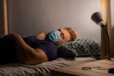 Kısa sarı saçlı genç çocuk yatağında yatıyor. Koronavirüse karşı koruyucu bir maskeyle.