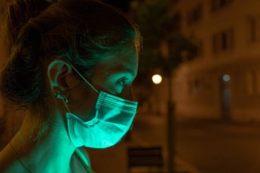 Gece sokakta yüzü aydınlanmış bir şekilde cep telefonunu kullanan genç İspanyol kadın Covid19 'a karşı maske takıyor. Arka planda kimse yok.