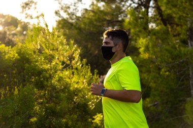 Siyah saçlı ve kahverengi tenli genç İspanyol bir adamın arka planda güneşle birlikte çam ormanında koşarken, Covid19 'a karşı maske taktığı ve sarı bir tişört giydiği yakın plan.
