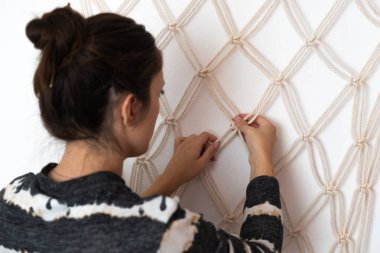 Genç İspanyol kız makrome perdenin düğümlerinin doğru olup olmadığını kontrol ediyor, beyaz duvarda asılı perde ve bej ip