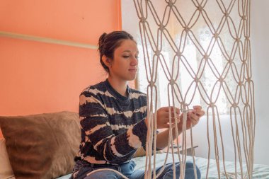 Yatak odasında makrome perde yapan İspanyol bir kızın arka planda parlak bir pencere ve beyaz bir perdeyle çekilmiş bir fotoğrafı..