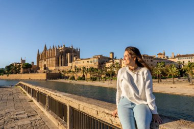 Palma katedralinin önünde oturan genç bir kadın altın gün batımını seyrediyor ve gülümsüyor, beyaz kazak ve çizgili pantolon giyiyor, açık gökyüzü ve küçük bir rüzgar.