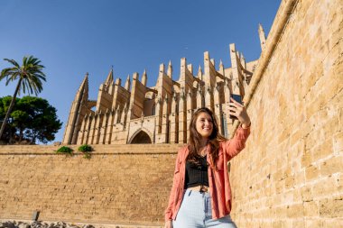 Açık tenli, kahverengi saçlı genç İspanyol kız öğleden sonra Palma de Mallorca katedralinde duvarın altından mutlu bir selfie çekiyor.