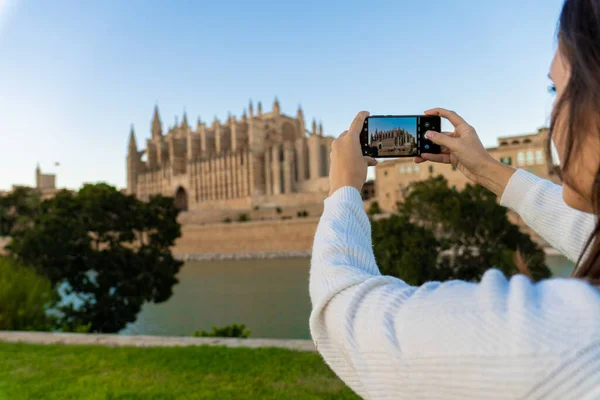 Young light-skinned girl taking a picture of the cathedral of Palma de Mallorca with her phone on her holidays.