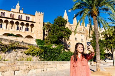 Açık tenli ve kahverengi saçlı genç İspanyol kız öğleden sonra Palma de Mallorca katedralinde arka planda insanlar olmadan selfie çekiyor.