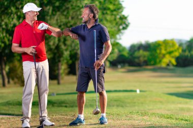 İki golfçü birbirini dirseklerine dokunarak selamlar. Golf arkadaşları o sırada merhaba der.