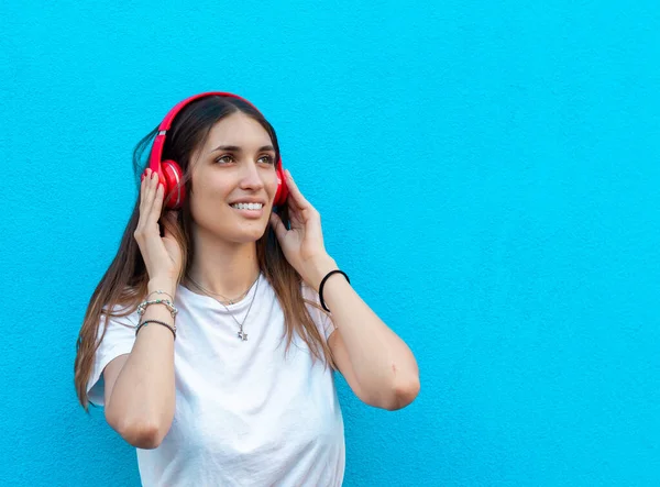 smiling young woman listen music with headphones in front of a blue wall - portrait of young girl with red headphones and blue blackground