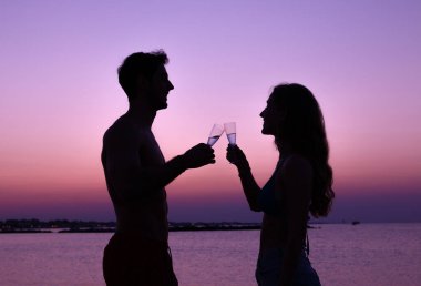 Gün batımındaki kumsalda aşık çift. Gün batımında kadeh kaldıran iki genç aşığın silueti.
