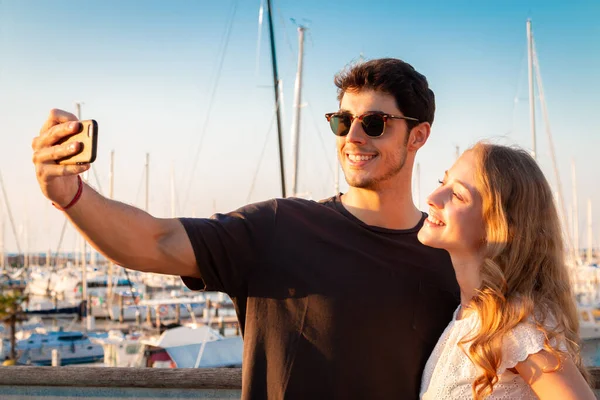 Genç çift gün batımında selfie çekiyor. İki genç turist turistik bir limanda selfie çekiyor.