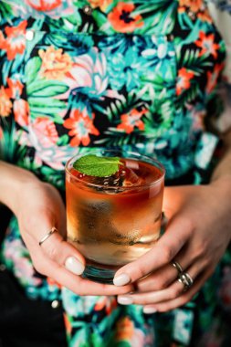 Elinde tiki tarzı renkli katmanlı bir kokteyl tutan bayan bardakta kristal berrak buz küpüyle, nane yaprağı ile süslenmiş.