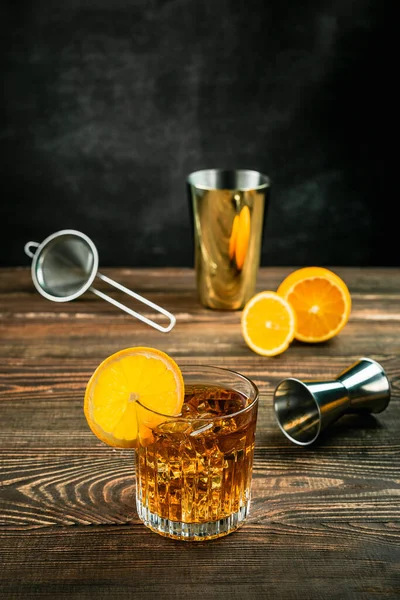 ロックグラスにオレンジアルコールカクテルをアイスで満たし オレンジホイールでガーニッシュ カットオレンジ ジッガー シェーカーとストレーナー 木製のテーブル 濃い灰色の背景 撮影所垂直ショット — ストック写真