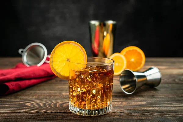 ロックグラスにオレンジアルコールカクテルをアイスで満たし オレンジホイールでガーニッシュ カットオレンジ ジッガー シェーカー ストレーナーと赤ナプキン 木製のテーブル 濃い灰色の背景 撮影所横ショット — ストック写真