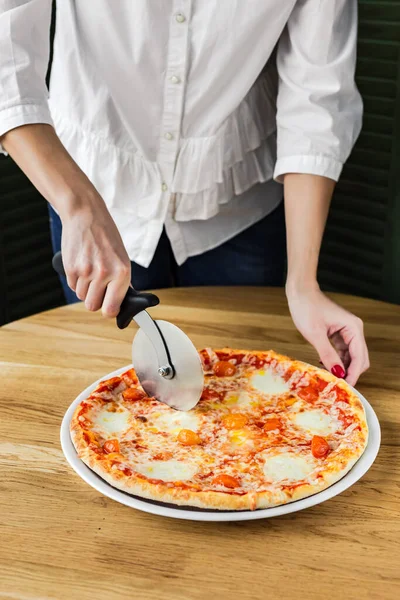 Cutting pizza margherita with a pizza cutter, served on a wooden round board, natural light