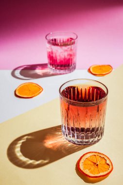 Kırmızı şuruplu iki buzlu bardağın çok parlak dikey fotoğrafı sert gölgeli sert ışık, turuncu cips. Pembe, beyaz ve sarı arkaplan
