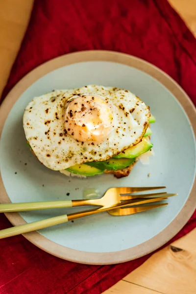 A rye-bread toast with cheese, avocado and fried egg for breakfast, served on a blue plate. Beautiful golden cutlery, red napkin.