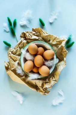 Paskalya 'da düz bir fotoğraf, bir kase kahverengi yumurtayı buruşuk el işi kağıdına, beyaz tüylere ve dekor olarak kuru yeşil bitkilere serer. Açık mavi arkaplan, yukarıdan görünüm