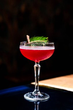 Coupe bardağında kırmızı ekşi bir kokteyl. Bardaki masanın üzerinde köknar dalı var.