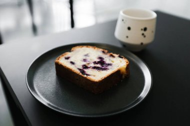 Bir parça meyveli kek, böğürtlenli ve siyah bir masada bir fincan kahve.