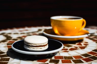Tabağında çikolata olan vanilyalı makarna, güneşli bir günde mozaik taş bir kafede sarı bir fincan sade kahve.