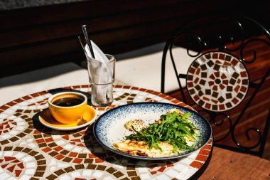 Güneşli bir günde bir kafenin mozaik taşı masasında roket salatası, bir fincan sade kahve ve roket salatasından oluşan bir kahvaltı.