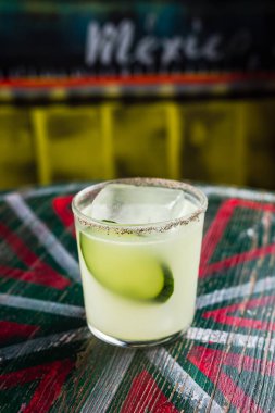 Büyük bir buzlu buzlu bardakta salatalık dilimiyle süslenmiş, cam kenarında baharatlı bir kokteyl.