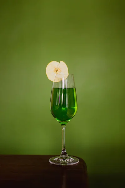A green cocktail in a flute glass garnished with a dehydrated apple slice on green background