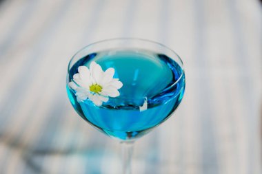 Çentikte parlak mavi bir kokteyl ve papatya çiçeğiyle süslenmiş Nora bardağı.