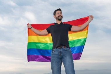 Elinde gay bayrağı olan bir adam orada oturmuş ufka bakıyor. Eşcinsel, gurur, çeşitlilik kavramı