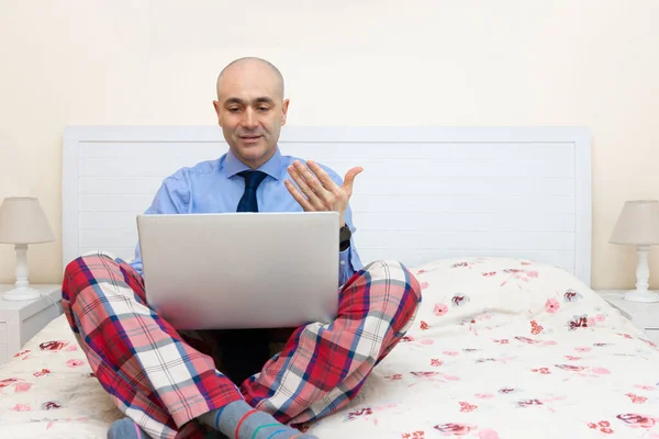 Yatakta dizüstü bilgisayarla çalışan bir adam gömlek, kravat ve pijama pantolon giyiyor. Uzaktan çalışma, açıklama ve uzaktan çalışma kavramı