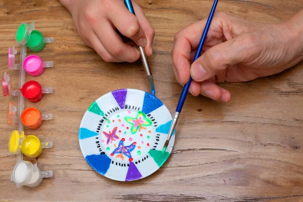 Aerial view of the hands of an adult and a child painting a porcelain plate with a brush and with a set of paints of different colors on top of wood. hobby concept