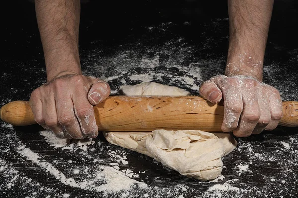 Hands of a man kneading a ball of flour with a wooden roller to make bread on a black surface full of flour