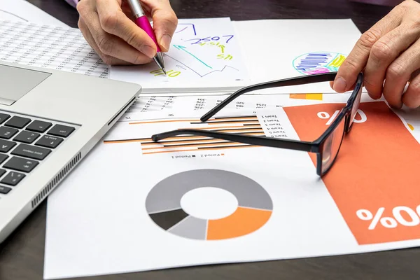 Detail of person hands drawing graphics on a paper and holding glasses with the other hand surrounded by a laptop and papers with numbers and graphics on a dark wooden desk