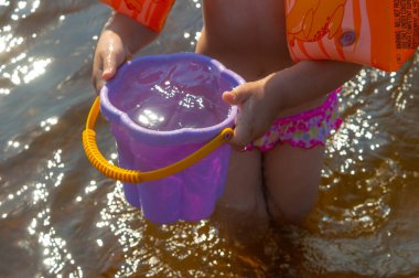 Turuncu kollu bir kız nehirde duruyor ve elinde mor bir kova su tutuyor.