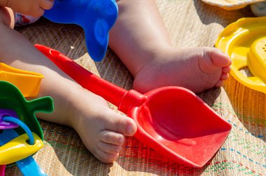 Çocuk kumsalda oyuncaklarla oturuyor. Çocuk kalıpları ve küreklerle oynuyor. Sadece bacakları görünüyor.