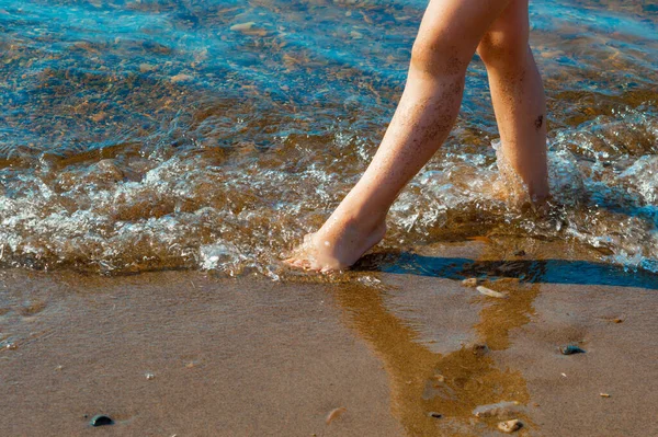 The child stands on the sandy bank of the river. Only legs are visible. River surf. Sunny summer day