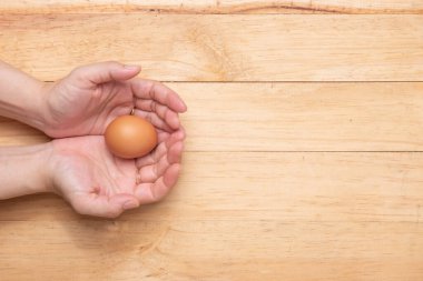 Açık kahverengi ahşap arka planda çiğ tavuk yumurtası tutan iki elin üst görüntüsü, boşluğu kopyala