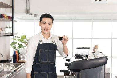 Gülümseyen Asyalı kahveci adam bir fincan sade kahve gösteriyor. Kahve dükkanındaki kameraya bakıp mutlu oluyor.