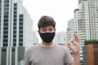 Asyalı adam, virüsü ve tozu korumak için siyah bir maske takıyor. El işareti tamam, COVID-19 konsepti.