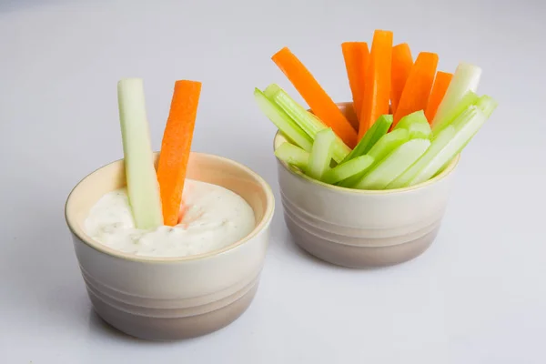 close up isolated shot of a bowl of crunchy orange carrot slices and juicy green celery sticks with a single carrot piece dipped in a white cup of blue cheese sauce on a white background