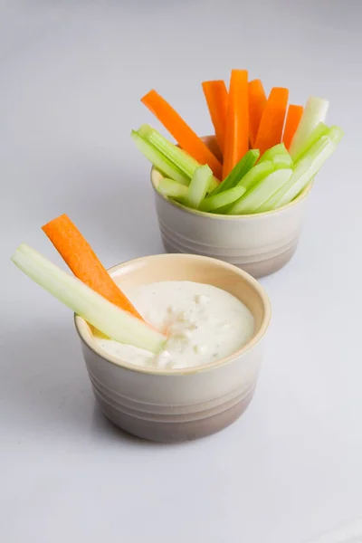 close up isolated shot of a bowl of crunchy orange carrot slices and juicy green celery sticks with a carrot and celery piece dipped in a white cup of blue cheese sauce on a white background