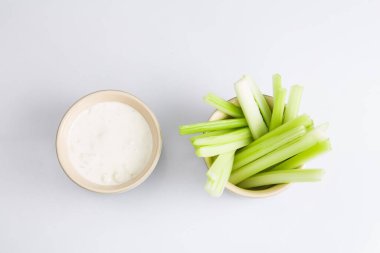 İzole edilmiş düz manzarayı kapatın. Beyaz arka planda beyaz bir kase mavi peynirli sosun yanında gevrek sulu yeşil kereviz çubuklarının üst görüntüsü yatıyordu.