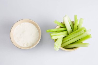 İzole edilmiş düz manzarayı kapatın. Beyaz arka planda beyaz bir kase mavi peynirli sosun yanında gevrek sulu yeşil kereviz çubuklarının üst görüntüsü yatıyordu.