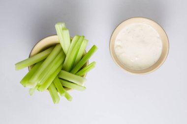 İzole edilmiş düz manzarayı kapatın. Beyaz arka planda beyaz bir kase mavi peynirli sosun yanında gevrek sulu yeşil kereviz çubuklarının üst görüntüsü yatıyordu.