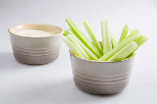 close up isolated top corner view shot of a bowl of crunchy juicy green celery sticks next to a white cup of blue cheese dipping sauce on a white background