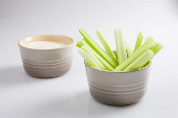 close up isolated top corner view shot of a bowl of crunchy juicy green celery sticks next to a white cup of blue cheese dipping sauce on a white background