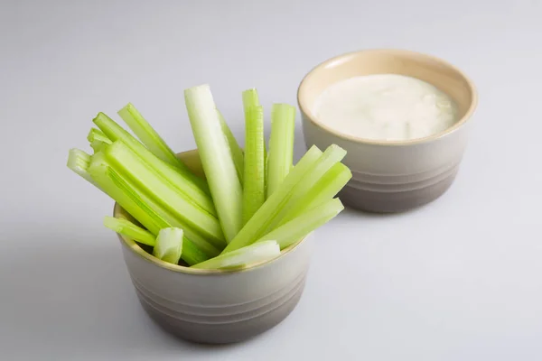 close up isolated top corner view shot of a bowl of crunchy juicy green celery sticks next to a white cup of blue cheese dipping sauce on a white background