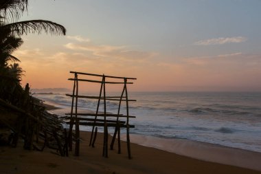 Güneş doğarken Pitiwella plajındaki (Sri Lanka) ahşap bir kemerin portakal kumu, yeşil palmiye ağaçları, kayalar, büyük beyaz dalgalı Hint okyanusu ve ufukta güneş olan güzel manzarası..
