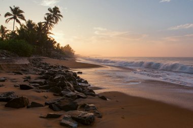 Pitiwella plajının (Sri Lanka) turuncu kum, yeşil palmiye ağaçları, kahverengi kayalar, büyük beyaz dalgalı Hint okyanusu ve ufukta güneş görünen güneş ile gün doğumunda çekilmiş güzel bir kartpostal manzarası..