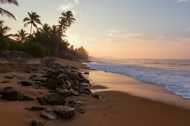 Pitiwella plajının (Sri Lanka) turuncu kum, yeşil palmiye ağaçları, kahverengi kayalar, büyük beyaz dalgalı Hint okyanusu ve ufukta güneş görünen güneş ile gün doğumunda çekilmiş güzel bir kartpostal manzarası..