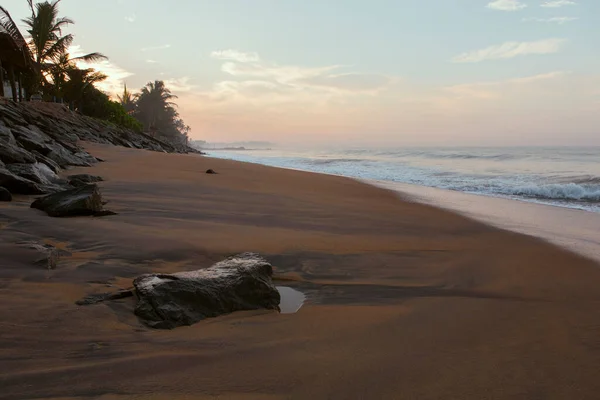 Pitiwella plajının (Sri Lanka) turuncu kum, yeşil palmiye ağaçları, kahverengi kayalar, büyük beyaz dalgalı Hint okyanusu ve ufukta güneş görünen güneş ile gün doğumunda çekilmiş güzel bir kartpostal manzarası.